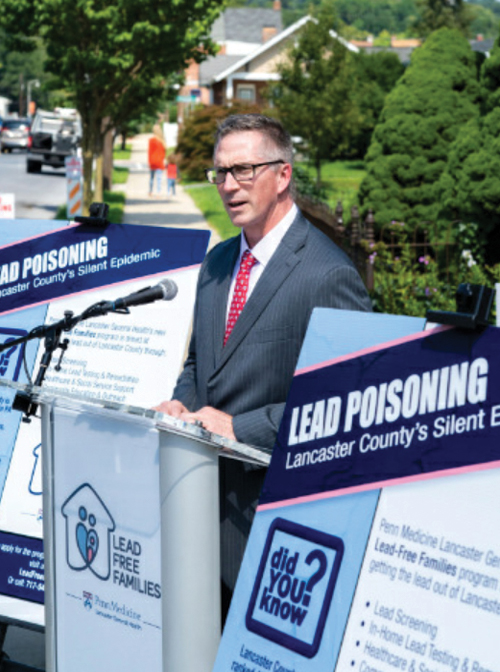 John J. Herman, CEO of Penn Medicine Lancaster General Health, stands outdoors behind a podium with Lead-Free Families posters on either side of him. 